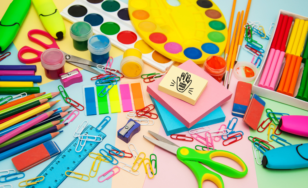 A table covered in an assortment of crafting supplies, such as watercolors, scissors, paperclips, rulers, highlighters, post it notes and erasers.  very colorful