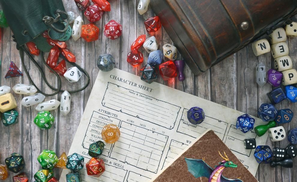 A wooden table top with a variety of dice spilling out across the table from all edges. There is an empty character sheet  and journal with the wing of a dragon showing at the bottom right corner.  A wooden box angles in from the top right corner with some letter cubes along that edge.