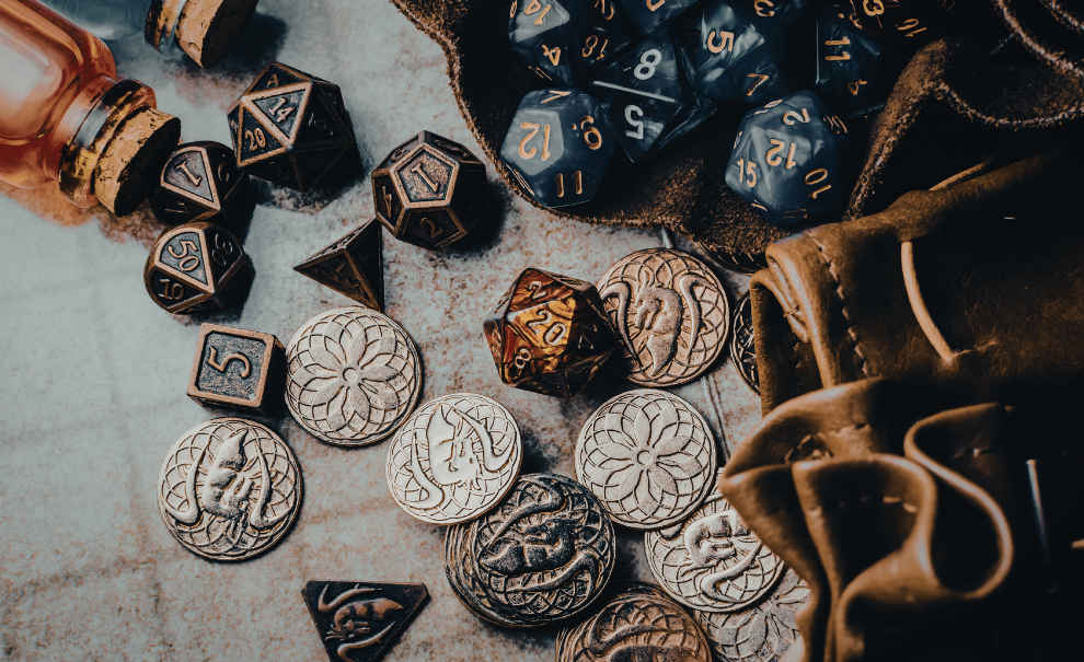 A group of metal polyhedral dice and fantasy coins. On the right hand side are two leather dice bags, the top one is open and full of blue plastic dice. There are two glass vials on the top left corner. They all sit on a gridded table. 