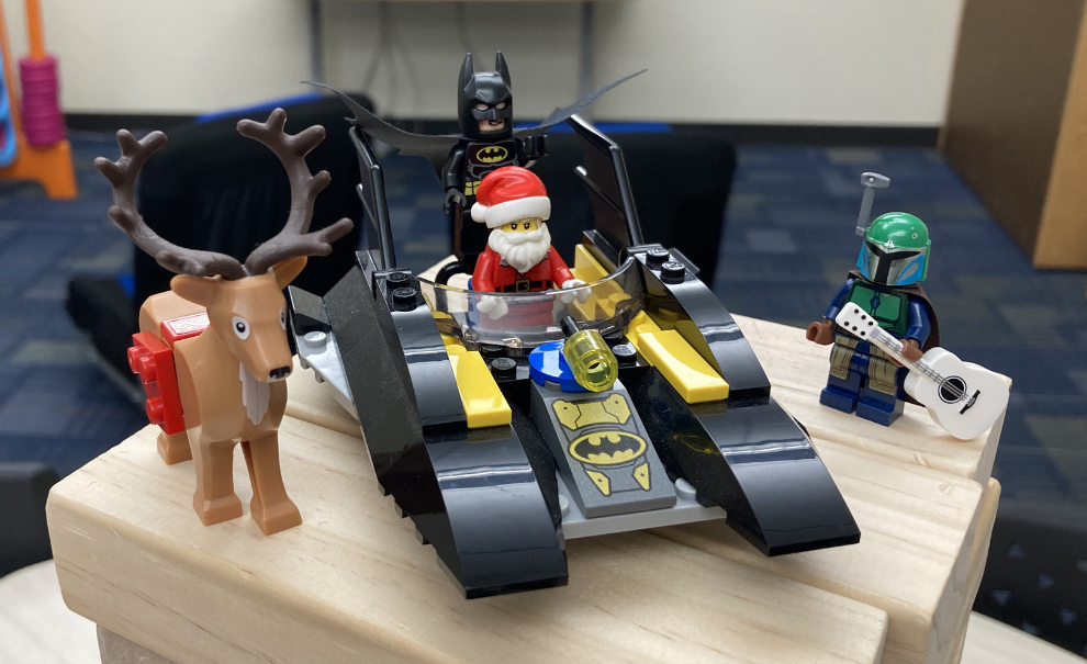 A LEGO Santa and Batman riding on a LEGO bat-boat. LEGO reindeer on their right and a LEGO Boba fett with a guitar on their left. 