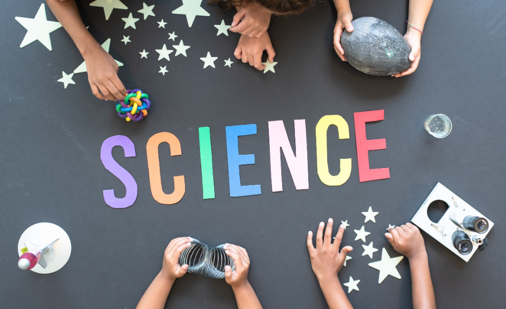 The word "Science" spelled out on a tabletop with several hands holding various science projects