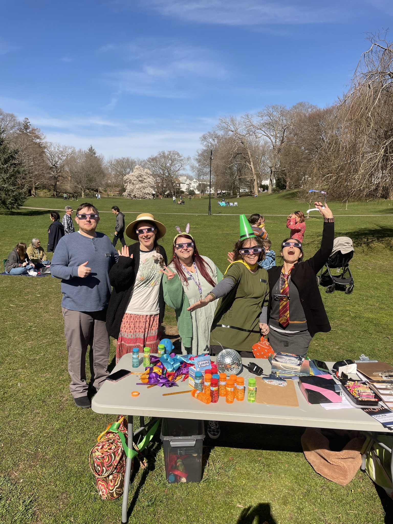 Photo of five Youth Services library staff in front of a table in Wilcox Park during the solar eclipse. Library workers are posing and waving with solar eclipse glasses on.