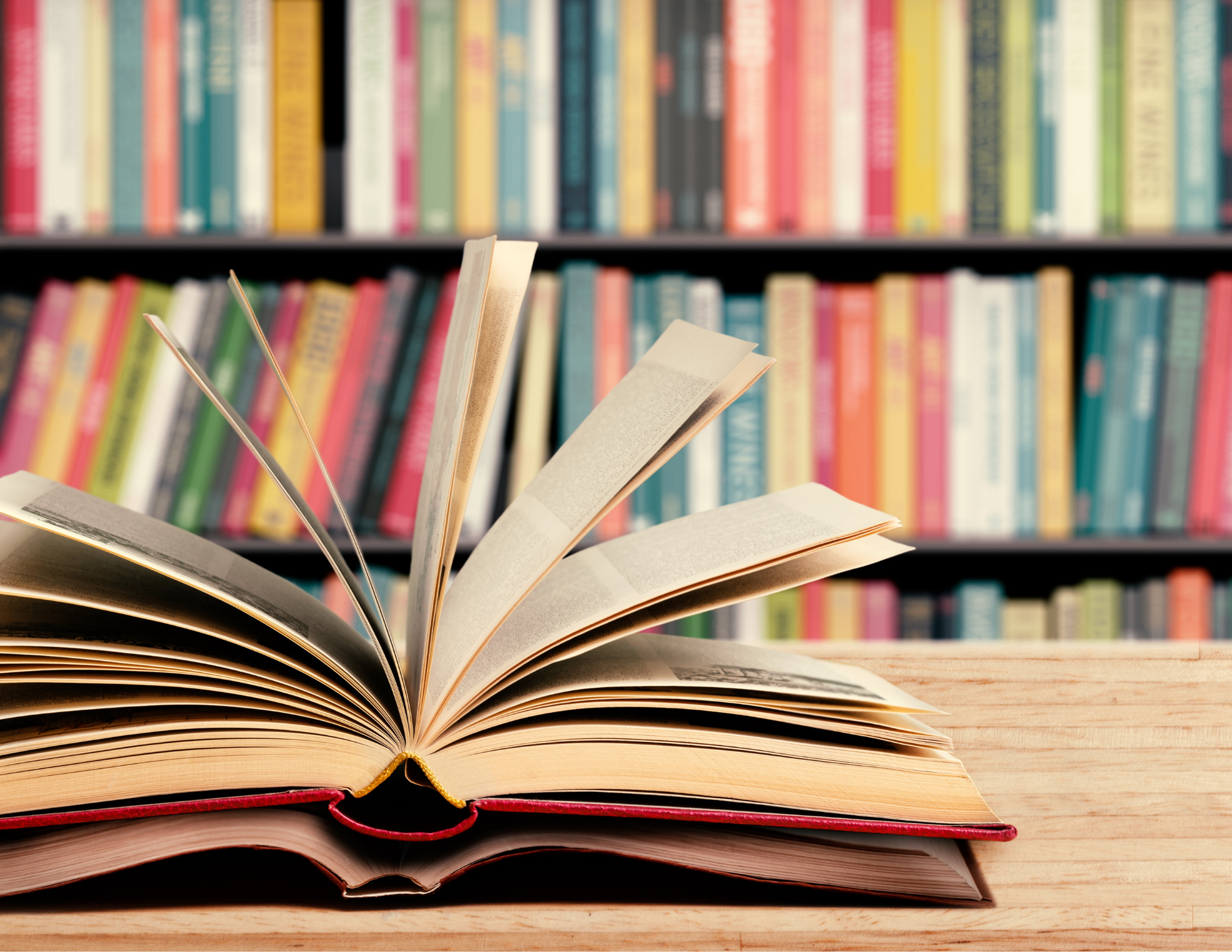 an open book with pages fanned out on a wooden table in the foreground with a bookcase full of colorful spines in the background.