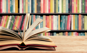 An open book laying on the left side of a wooden table with shelf full of books in the background. 