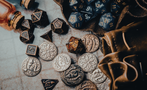 A group of metal polyhedral dice and fantasy coins. On the right hand side are two leather dice bags, the top one is open and full of blue plastic dice. There are two glass vials on the top left corner. They all sit on a gridded table. 