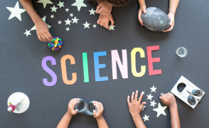 The word "Science" spelled out on a tabletop with several hands holding various science projects