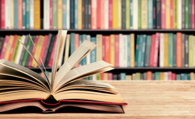 An open book laying on the left side of a wooden table with shelf full of books in the background. 