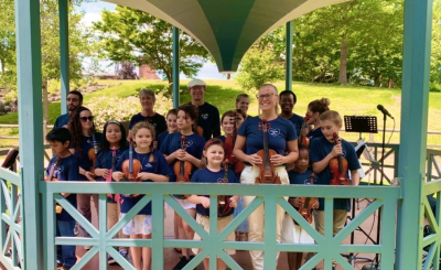 Group of kids and adult woman instructor with violins in a bandshell out in the park