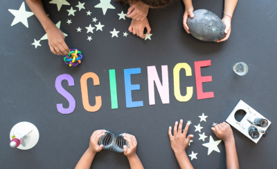 The word "Science" spelled out on a tabletop with several hands holding various science projects